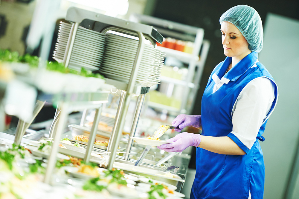 Person wearing hygiene hair net serving food on a plate in a canteen environment