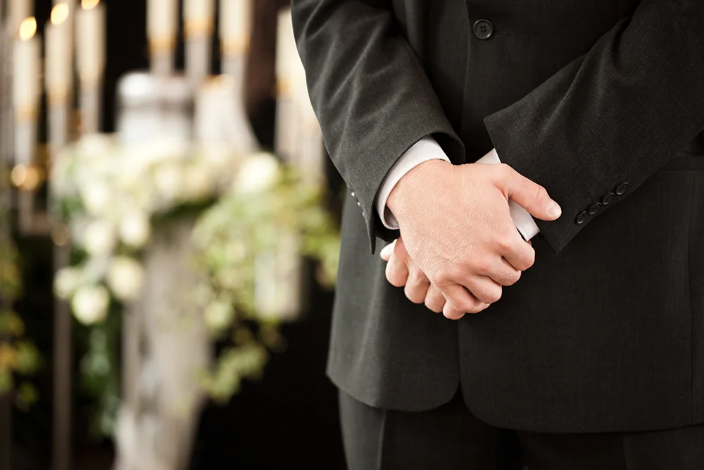 Close up of man's hands wearing black with funeral flowers in the background