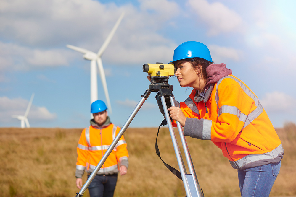 Person wearing helmet and high visability jacket using a surveying camera out in a field. 